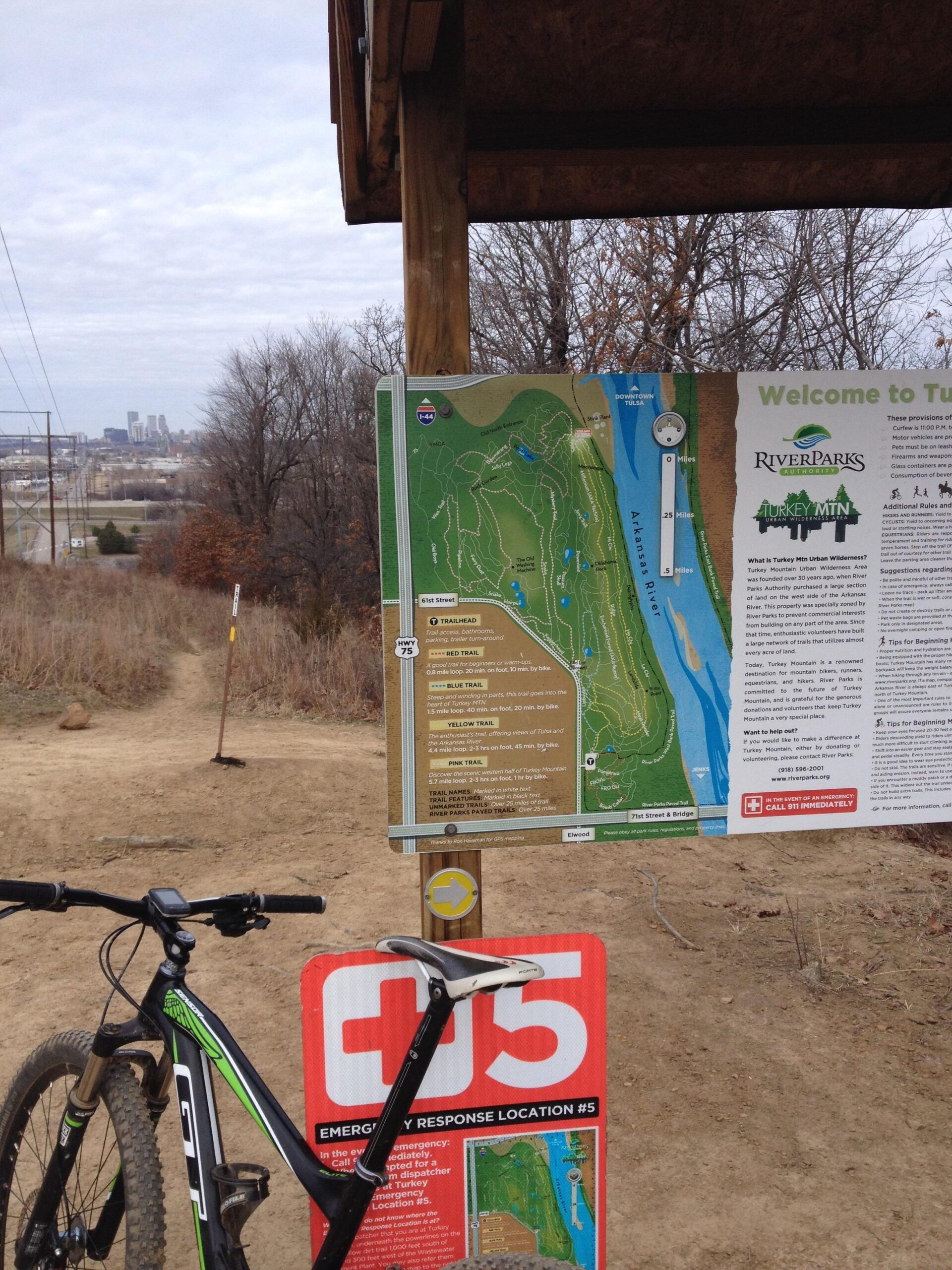 An outdoor scene featuring a mountain biking trail information board at Turkey Mountain Urban Wilderness Area. The board displays a detailed map of the trails, including colored routes for different difficulty levels, alongside emergency response location signage. A black and green mountain bike is positioned in the foreground with a view of the city skyline in the distance, under a cloudy sky. Turkey Mountain mountain bike trail.