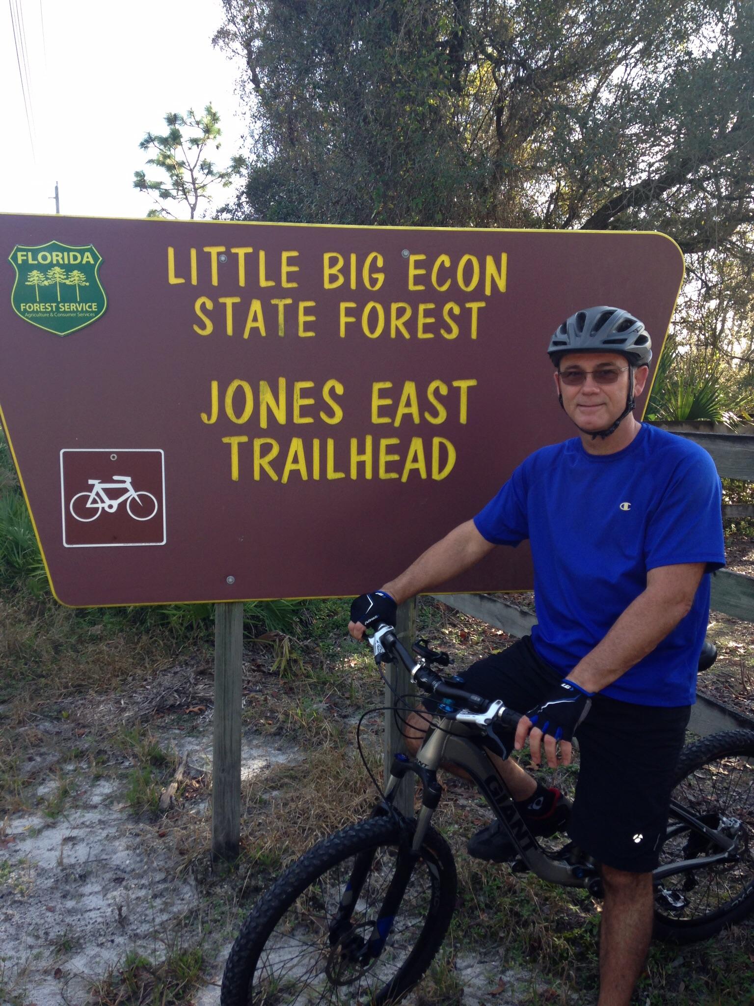 A person wearing a blue shirt and cycling gloves sits on a mountain bike next to a trailhead sign for the Little Big Econ State Forest, marked as "Jones East Trailhead." The sign features forest service branding and indicates that the area is suitable for biking. Surrounding vegetation and a clear sky are visible in the background. Little Big Econ State Forest mountain bike trail.