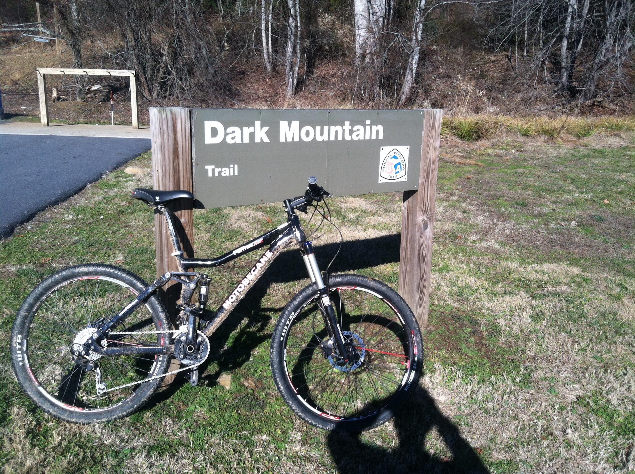 A mountain bike resting next to a sign that reads "Dark Mountain Trail," with green grass and trees in the background. The sign indicates the start of a biking trail, suggesting an outdoor recreational area. Dark Mountain Trail mountain bike trail.