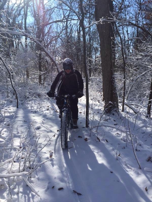 A person riding a mountain bike through a snow-covered wooded path on a clear, sunny day. Snow blankets the ground and clings to the branches of the trees, creating a winter wonderland. The cyclist is dressed in warm clothing and a helmet, navigating the serene, snowy landscape. Rock Cut State Park mountain bike trail.