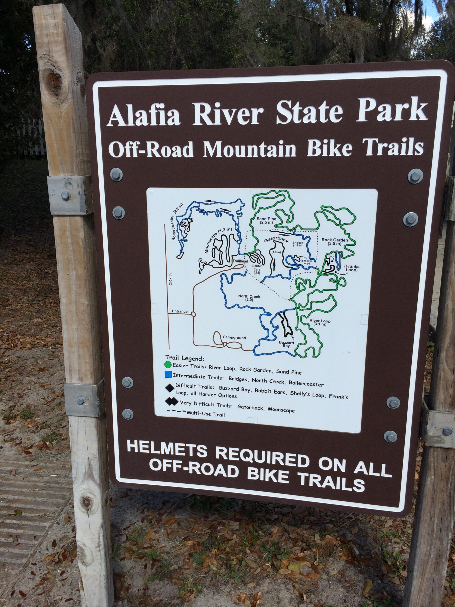 Sign for Alafia River State Park displaying a map of off-road mountain bike trails, including trail types and lengths. Text at the bottom states that helmets are required on all off-road bike trails. The sign is mounted on a wooden post in a natural setting. Alafia River State Park mountain bike trail.