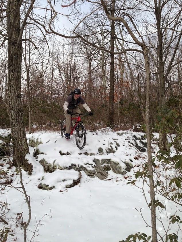 A mountain biker is descending a rocky ledge covered in snow, surrounded by trees in a winter forest setting. The rider is wearing a helmet and is in a dynamic pose, balancing on the bike as he navigates the snowy terrain. Frederick Watershed mountain bike trail.