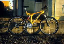GT xcr-3000: A brightly colored yellow mountain bike positioned on a gravel surface beside a building. The bike features a sturdy frame, wide tires, and a suspension system, highlighting its suitability for off-road riding.