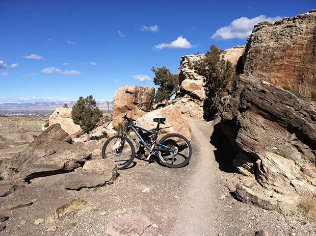 Alt text: A mountain bike is parked on a narrow dirt trail surrounded by rocky formations and sparse vegetation under a clear blue sky. The landscape features large boulders and distant hills.
