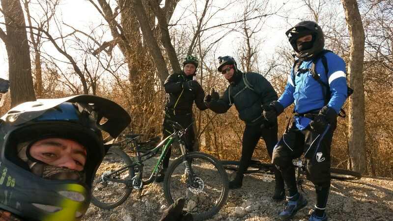 Four mountain bikers posing outdoors, wearing helmets and protective gear, with a bicycle resting nearby. The background features bare trees and a rocky terrain, suggesting a winter or early spring setting. One biker is taking a selfie, creating a lively group shot. Palos Forest Preserve mountain bike trail.
