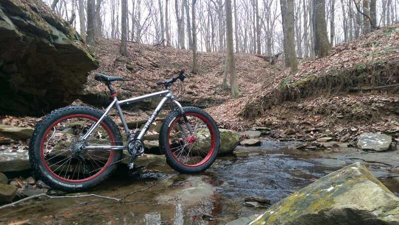 A silver mountain bike with red accents parked next to a shallow stream in a wooded area, surrounded by bare trees and fallen leaves. The rocky terrain and tranquil setting suggest a natural environment ideal for outdoor biking adventures. West Branch mountain bike trail.