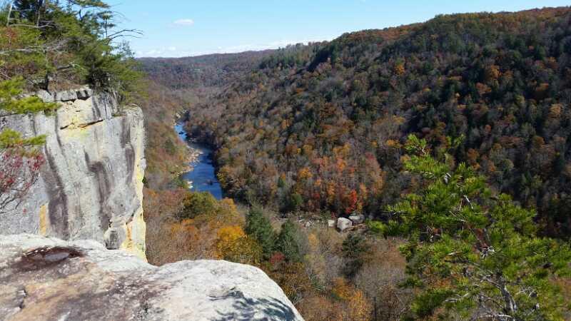 A scenic view from a rocky cliff overlooking a river winding through a valley filled with autumn foliage. The landscape features a mix of green and orange trees, with a clear blue sky in the background. Grand Gap mountain bike trail.