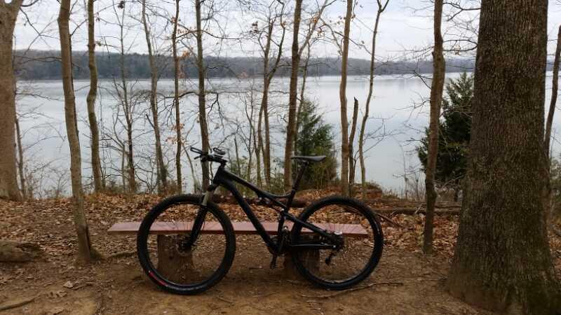 A black mountain bike is positioned on a wooden bench near a serene lake surrounded by bare trees, with a cloudy sky overhead. The scene reflects a peaceful outdoor setting ideal for recreational activities. Lock 4 mountain bike trail.