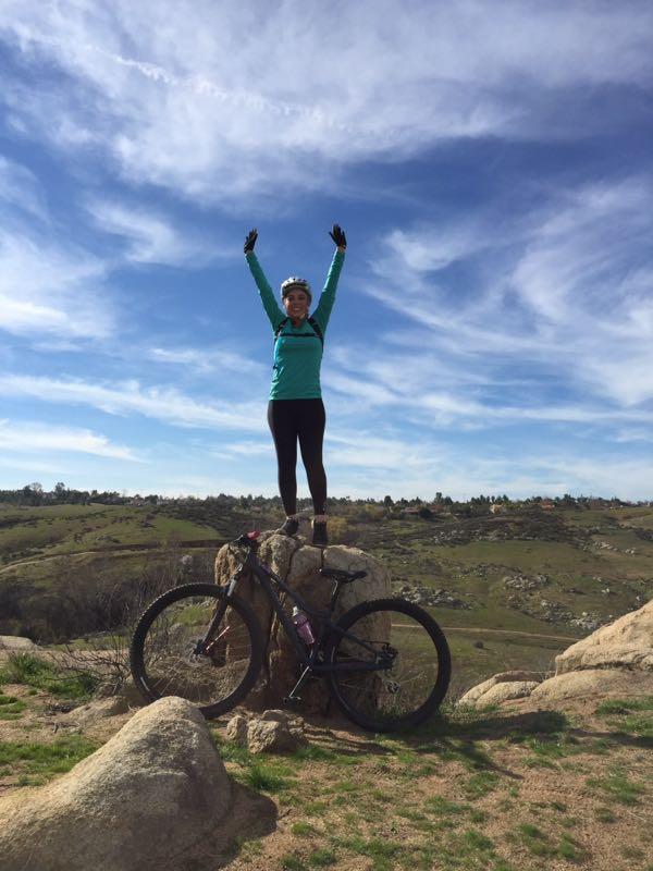 A person wearing a turquoise jacket and a helmet stands triumphantly on a rock with arms raised, next to a mountain bike. The backdrop features a scenic landscape of rolling hills under a blue sky with wispy clouds. Sycamore Canyon Park mountain bike trail.