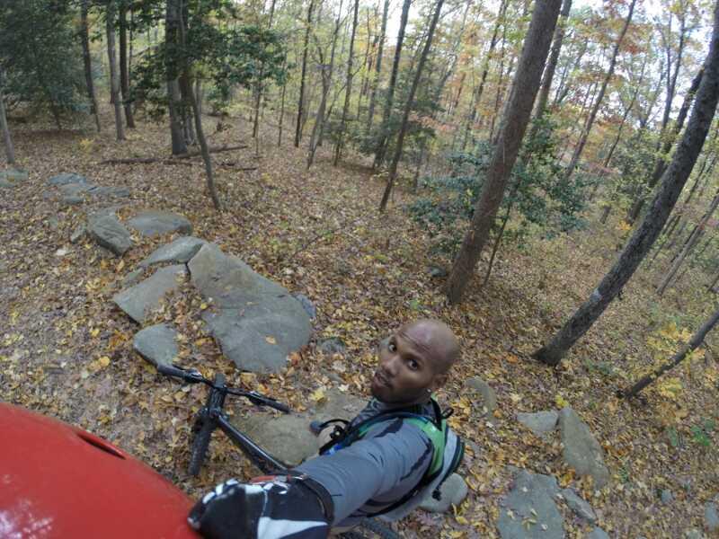 A person taking a selfie while mountain biking on a rocky trail in a forest with autumn foliage. The surroundings feature trees with changing leaves and scattered rocks on the ground. Powhite Park mountain bike trail.