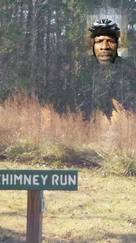 A wooden sign labeled "Chimney Run" stands in a grassy area with tall, brown grass and trees in the background. In the upper part of the image, a person wearing a bicycle helmet appears, partially blended with the scenery. Southwest Park mountain bike trail.
