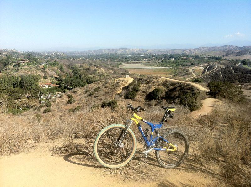 A mountain bike rests on a trail overlooking a scenic valley with rolling hills, greenery, and a winding river. The sky is clear with a few clouds in the distance, showcasing a beautiful outdoor landscape. Peters Canyon Regional Park mountain bike trail.