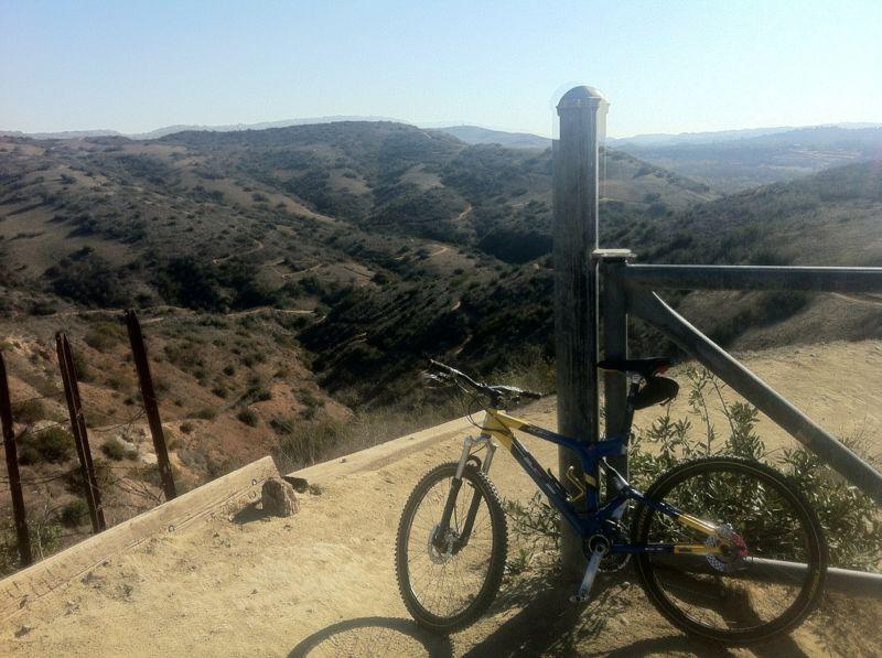 A mountain bike rests against a wooden post at a scenic overlook, with rolling hills and valleys visible in the background under a clear sky. Santiago Oaks mountain bike trail.