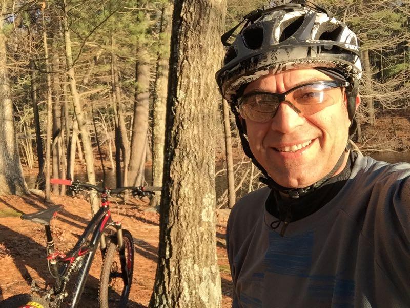 A person wearing a cycling helmet and sunglasses smiles for a selfie beside a mountain bike parked against a tree in a forested area. The background features tall trees and a hint of sunlight filtering through the foliage, suggesting a pleasant outdoor environment. Highland Mountain Bike Park mountain bike trail.