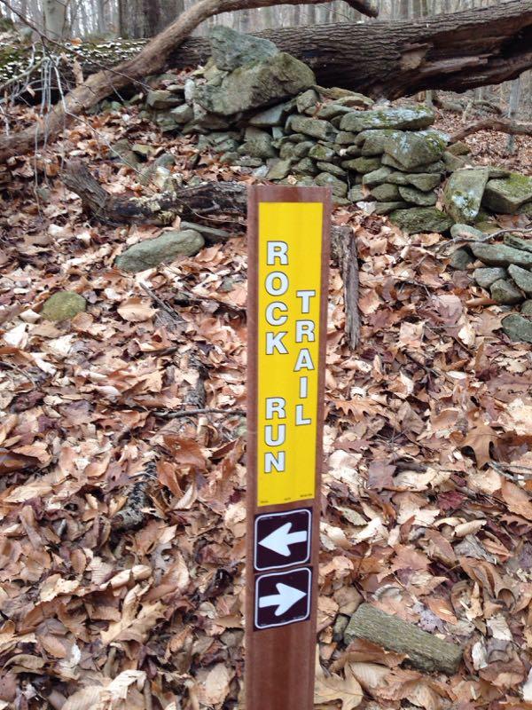 Trail sign indicating "Rock Run Trail," with arrows pointing left and right, surrounded by fallen leaves and a stone wall in the background. Susquehanna mountain bike trail.