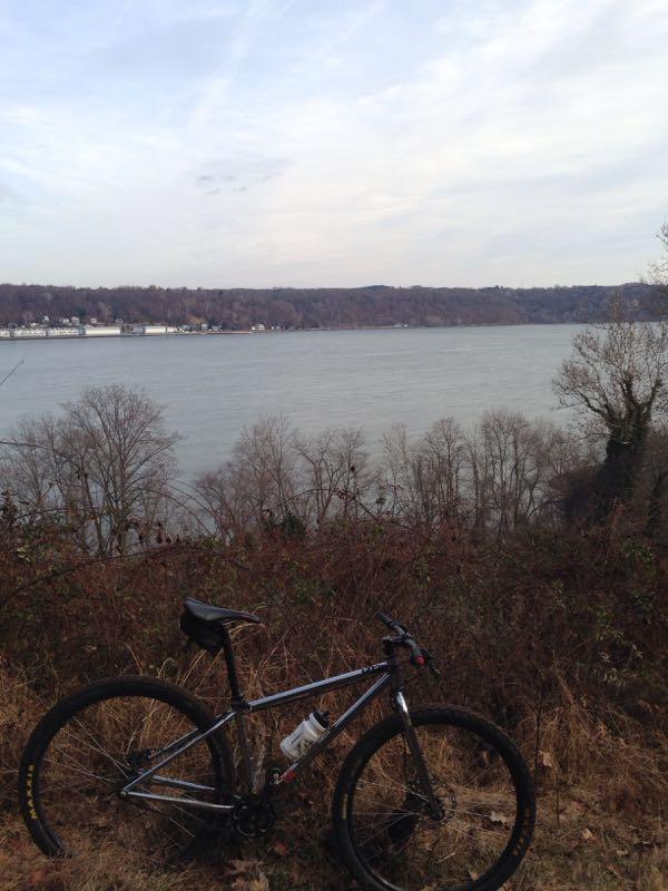 A mountain bike resting on a hillside overlooking a calm river. The landscape is framed by bare trees and distant hills, with a cloudy sky above. Susquehanna mountain bike trail.
