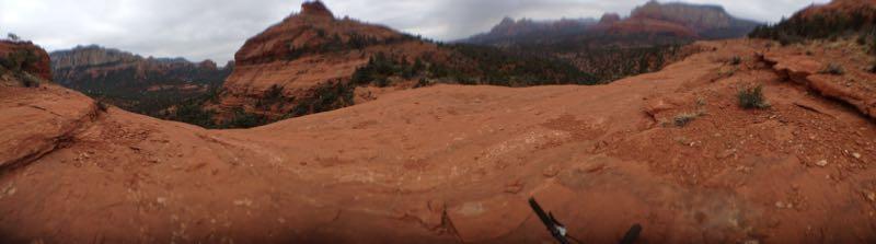 A panoramic view of a rugged red rock landscape, featuring a wide, flat terrain with scattered vegetation, surrounded by towering rock formations and distant mountains under a cloudy sky. Hangover mountain bike trail.