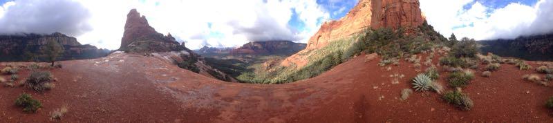 A panoramic view of a rugged landscape featuring red rock formations, green vegetation, and a cloudy blue sky. The foreground shows a reddish soil area with sparse plants, leading to steep cliffs and mountains in the background. Hangover mountain bike trail.