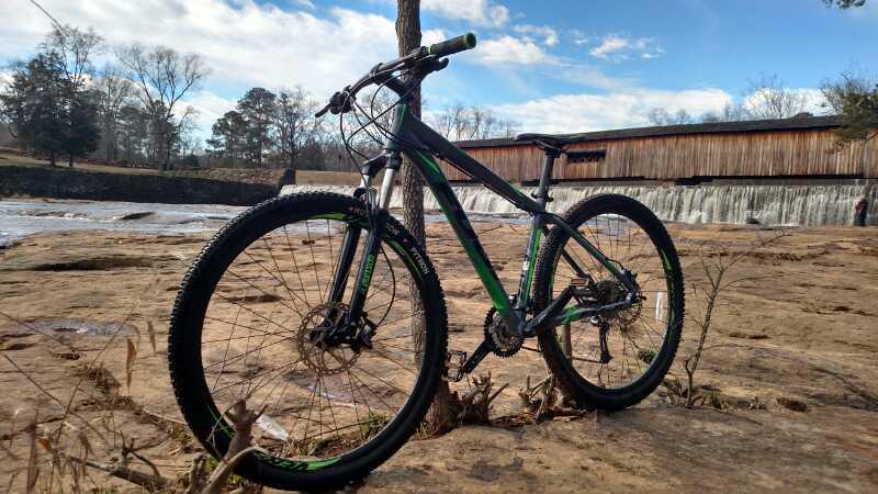 A mountain bike on rocky terrain with a waterfall in the background and clear blue sky, showcasing the bike's green and black design. Watson Mill Bridge State Park mountain bike trail.