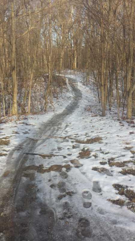 A narrow, winding dirt path lined with trees, partially covered in snow and ice, leading through a winter forest. Footprints are visible in the snow along the path. Alum Creek Phase I mountain bike trail.
