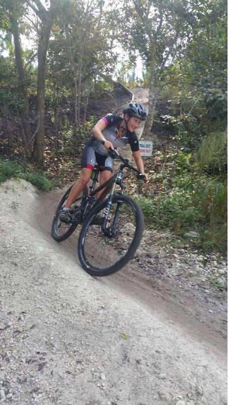 A person in athletic cycling gear riding a mountain bike down a dirt trail surrounded by trees. The trail is slightly sloped, with a sign visible in the background. The cyclist is focused on maintaining balance as they navigate the terrain. Markham Park mountain bike trail.