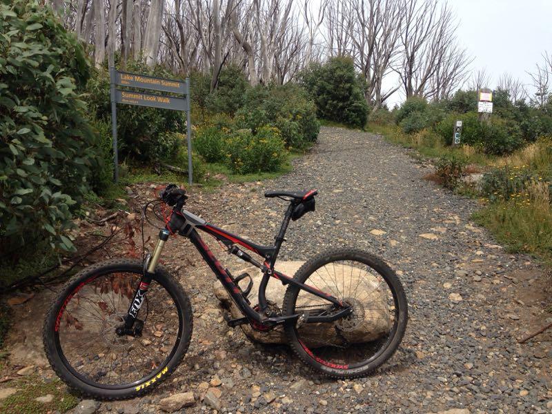 A mountain bike resting on a smooth rock alongside a gravel path leading to the Lake Mountain Summit trail. In the background, a sign indicates the direction to the Lake Mountain Summit and Summit Look Walk. Surrounding vegetation includes shrubs and small flowers, with a backdrop of bare trees. Lake Mountain mountain bike trail.