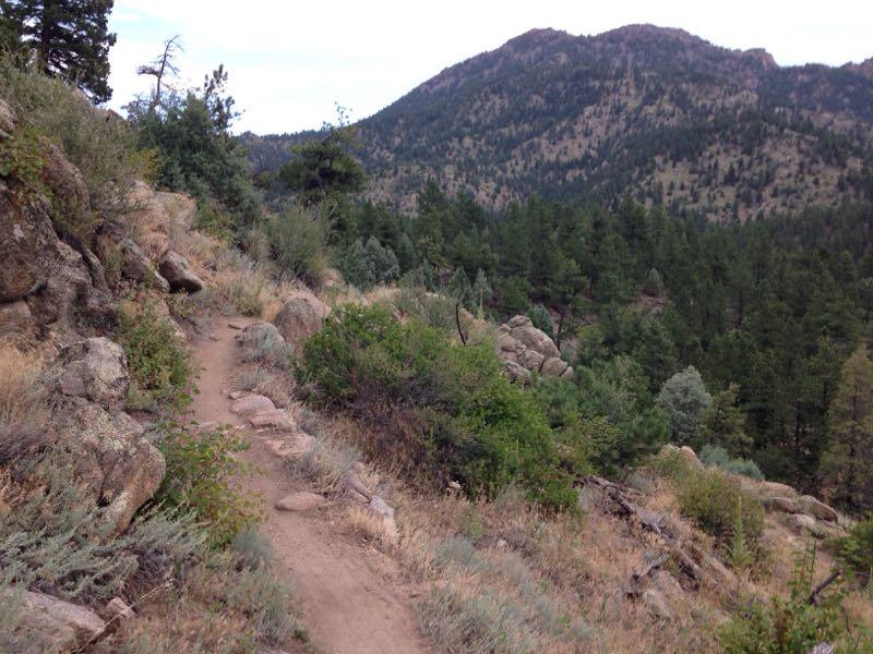 A winding dirt trail runs alongside rocky outcrops and lush greenery, leading towards a backdrop of rolling mountains under a partly cloudy sky. Dense trees and shrubs line the path, creating a natural, serene landscape. Walker Ranch mountain bike trail.