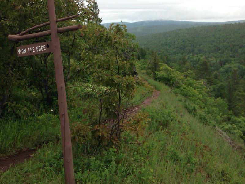 A wooden signpost labeled "On the Edge" stands along a grassy trail overlooking a vast landscape of lush green hills and mountains under a cloudy sky. Copper Harbor Trails mountain bike trail.