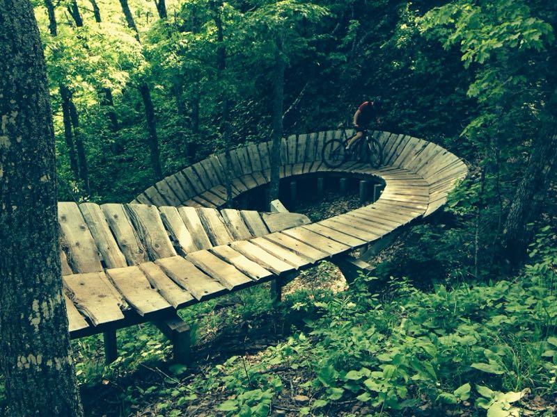 A mountain biker riding on a wooden, winding bike path through a lush green forest. The path is elevated and curves smoothly, surrounded by trees and foliage. Copper Harbor Trails mountain bike trail.