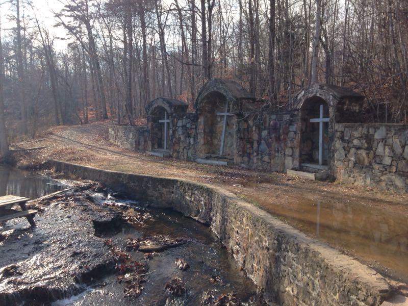 A tranquil scene of a stone wall with three arches, each featuring a white cross, nestled among bare trees. In the foreground, a gently flowing stream runs alongside a pathway that leads into the wooded area, capturing the serene atmosphere of the natural surroundings. Liberty Mountain mountain bike trail.