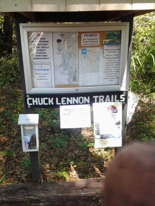 Signage for Chuck Lennon Trails, featuring a trail map, safety information, and guidelines for helmet usage. The sign is placed under a shelter and includes flyers for local events and trail updates. Surrounding vegetation is visible, indicating a natural setting. Chuck Lennon Park mountain bike trail.