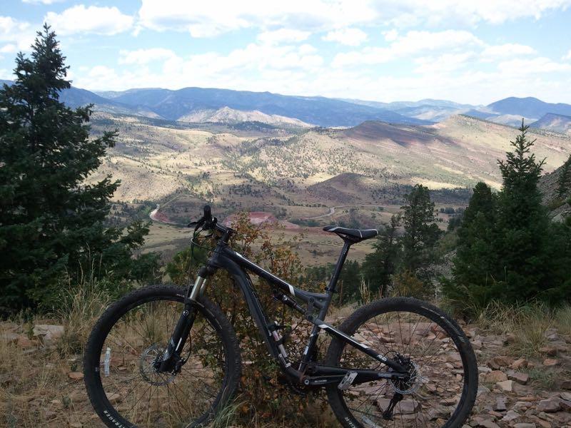 A mountain bike positioned on a rocky trail, overlooking a vast mountainous landscape with rolling hills and scattered trees. The sky is partly cloudy, adding to the scenic beauty of the outdoor setting. Heil Valley Ranch mountain bike trail.