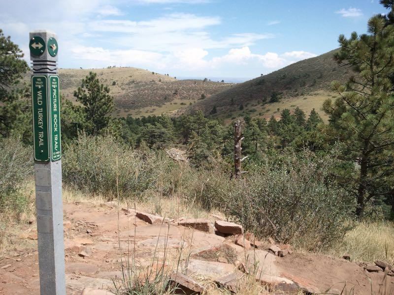 A trail sign marking the Wild Turkey Trail and Picture Rock Trail, with a scenic view of rolling hills and a forested area beneath a partly cloudy sky. Heil Valley Ranch mountain bike trail.