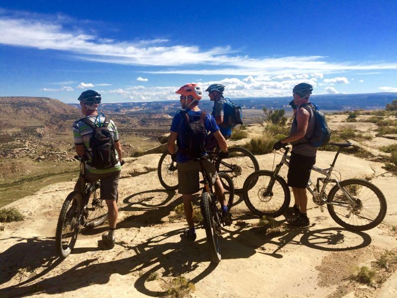 Four mountain bikers stand on a rocky outcrop, overlooking a vast landscape under a clear blue sky. They are wearing helmets and backpacks, with their bikes positioned beside them. The scenery features rolling hills and distant mesas, creating a picturesque backdrop for outdoor adventure. Western Rim mountain bike trail.