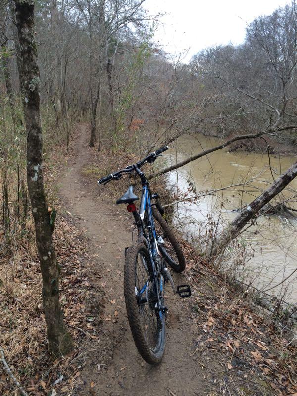 A mountain bike parked on a narrow dirt trail alongside a river, surrounded by bare trees and foliage, on a cloudy day. Rocky River Trail mountain bike trail.
