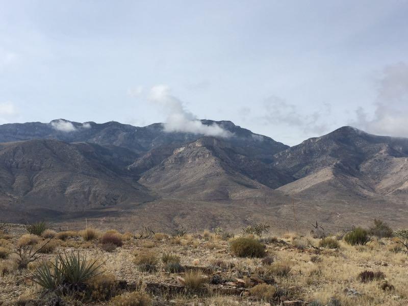 A panoramic view of rugged mountains under a cloudy sky, featuring layers of rocky terrain and sparse vegetation in the foreground. The landscape reflects a desert-like environment with varying shades of brown and green. Badger Pass Loop mountain bike trail.
