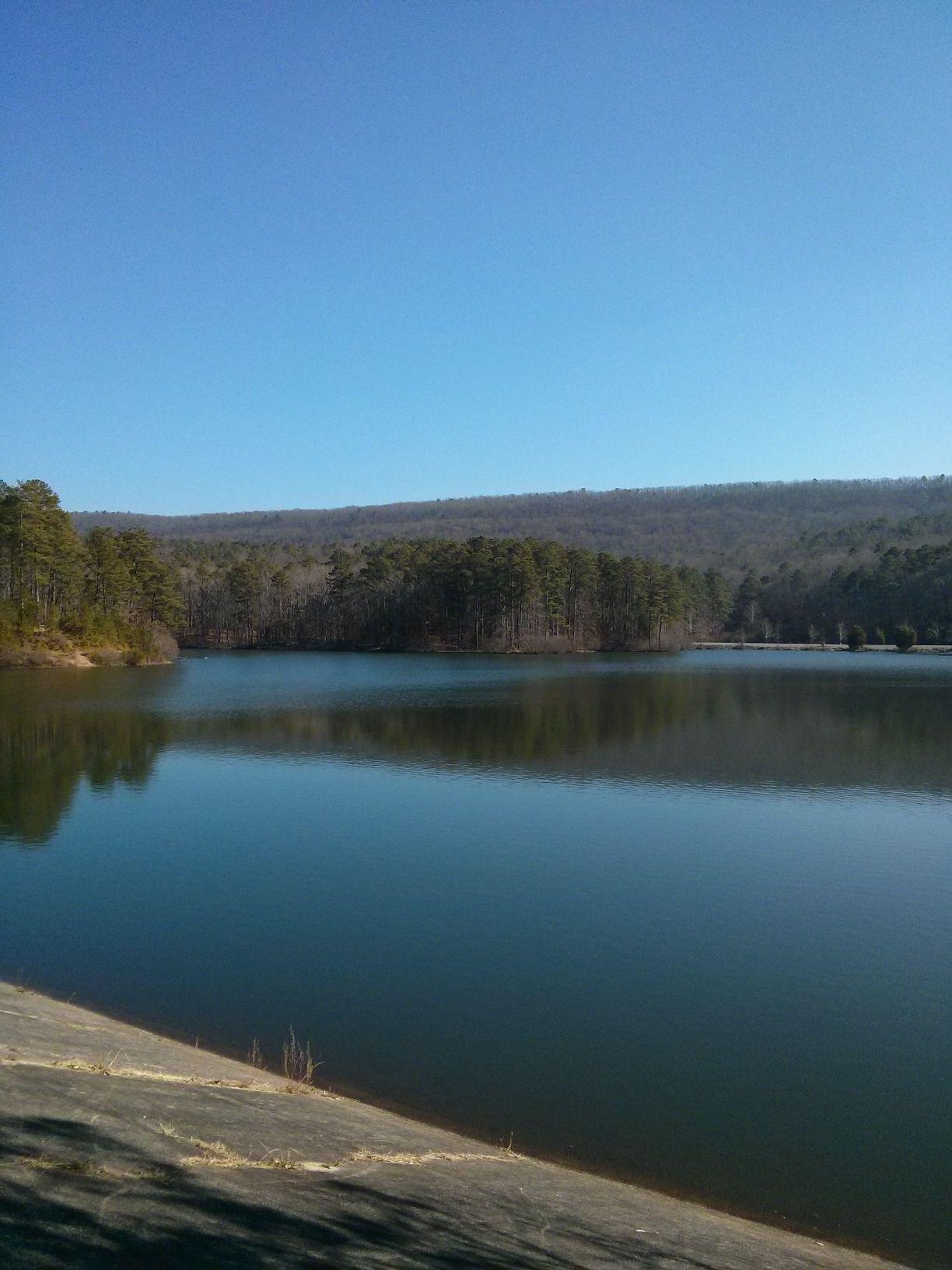 A serene landscape featuring a calm lake surrounded by lush green trees and a distant rolling hillside under a clear blue sky. The still water reflects the natural surroundings, creating a peaceful atmosphere. Oak Mountain State Park Bump Trail mountain bike trail.