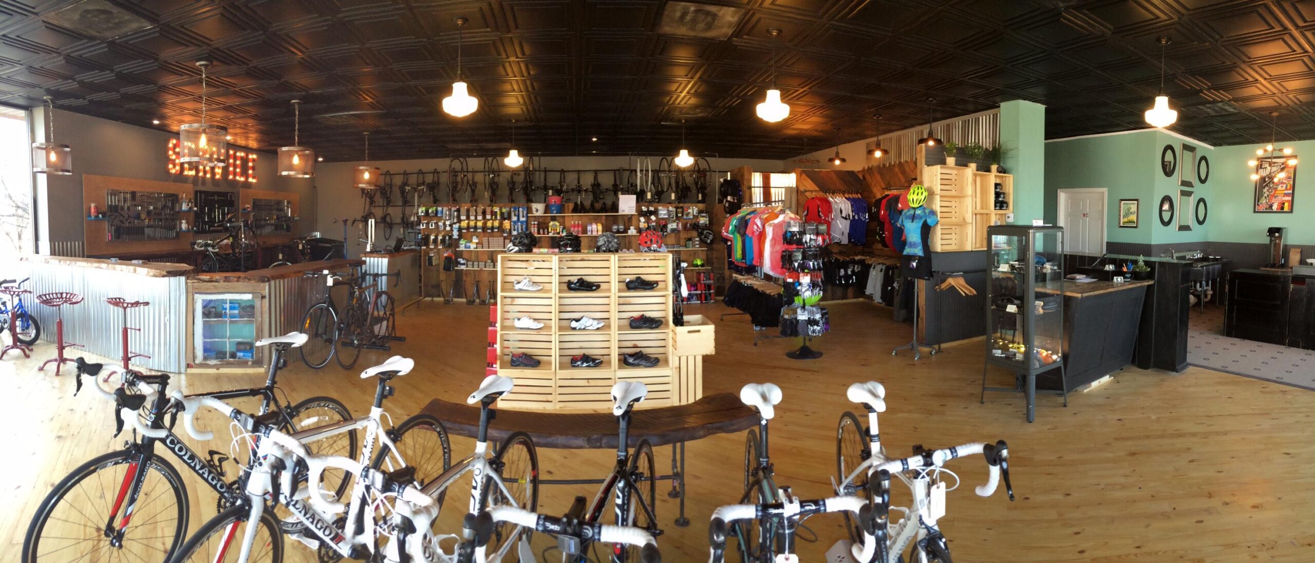Alt text: A brightly lit bicycle store featuring a variety of bikes displayed prominently in the foreground. The interior has wooden flooring and a stylish black ceiling with hanging lights. Shelves stocked with cycling gear, clothing, and accessories line the walls, while a service area with tools can be seen in the background.