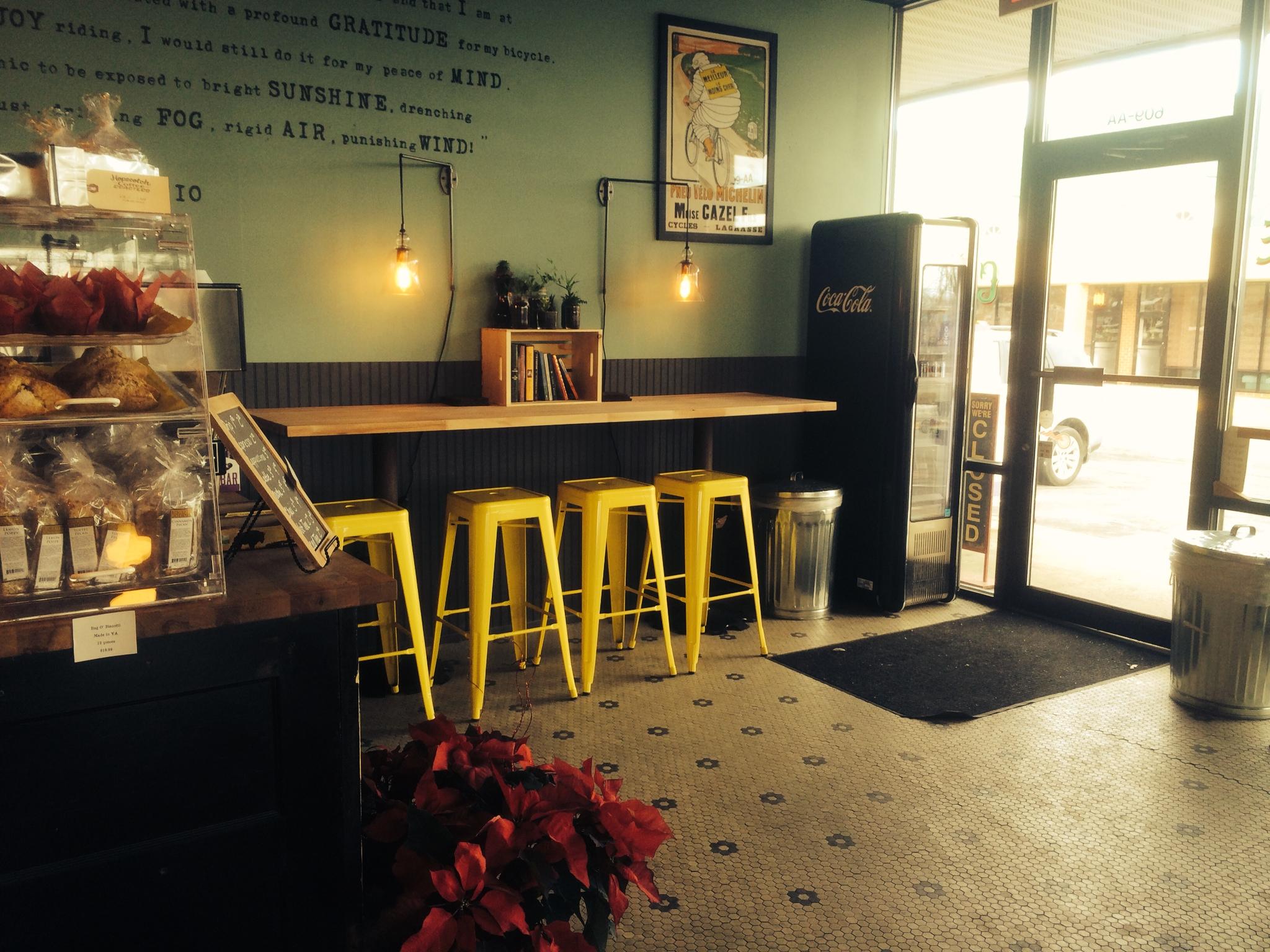 A cozy café interior featuring a display of pastries, a long wooden countertop with yellow stools, and a Coca-Cola fridge. The walls are adorned with decorative light fixtures and a poster, while potted plants add a touch of greenery. The entrance is visible with a "Closed" sign, and the floor is patterned with black and white tiles.