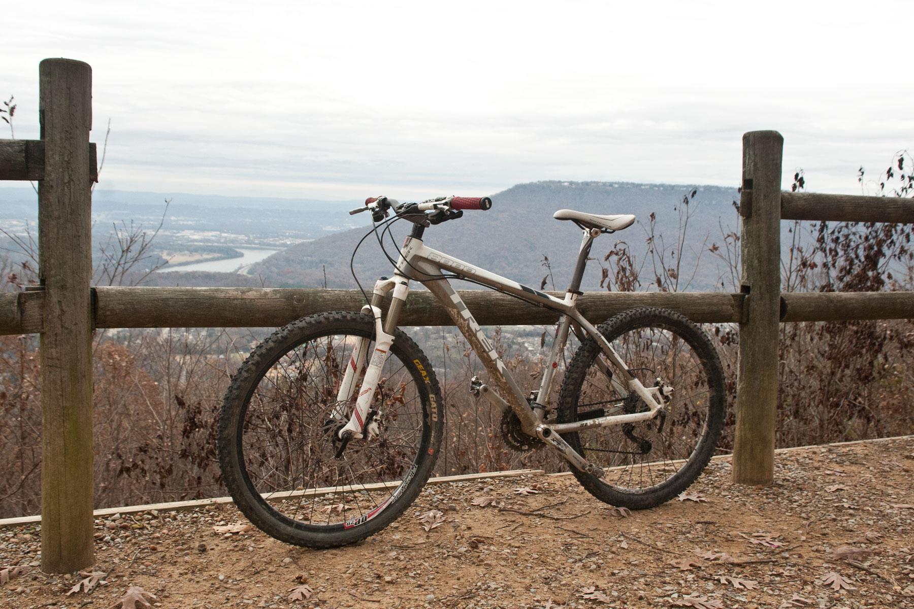 Trek 8000: A mountain bike parked on a dirt path beside a wooden fence, overlooking a scenic valley with a river. The landscape features distant hills and a cloudy sky, indicating a cool, overcast day. Dry leaves are scattered on the ground.