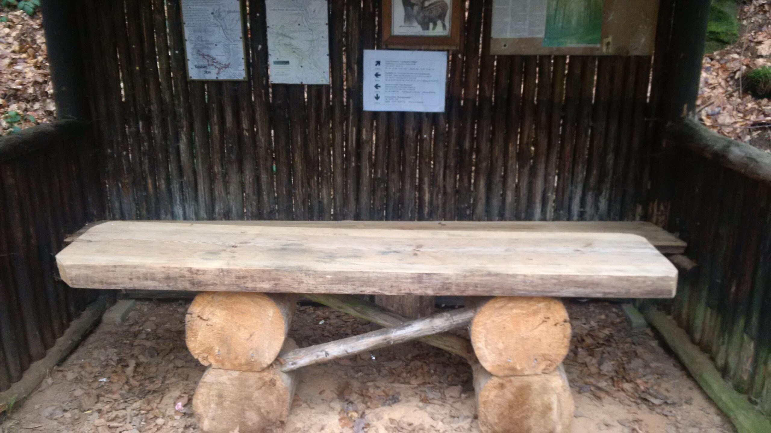 A rustic wooden bench made from logs, located inside a simple wooden shelter. The background features wooden walls and various informational posters mounted on the back wall. Leaves and dirt cover the ground inside the shelter, suggesting a natural outdoor setting. Countess Sonja Bernadotte Way mountain bike trail.