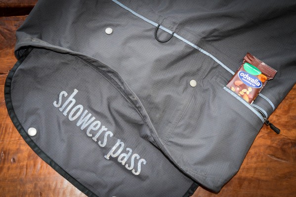 A close-up of a black waterproof jacket featuring the brand name "showers pass" embroidered on the back. The jacket has a zipper pocket containing a bar of Odwalla chocolate-covered nuts, resting on a wooden surface.