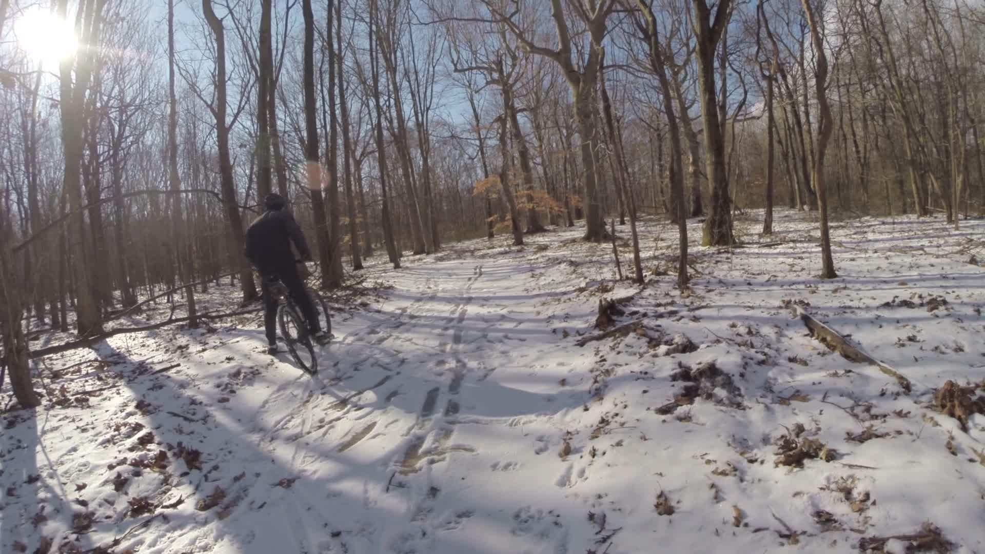 A person riding a mountain bike on a snow-covered trail in a wooded area, with bare trees and sunlight filtering through the branches. Long Pond mountain bike trail.