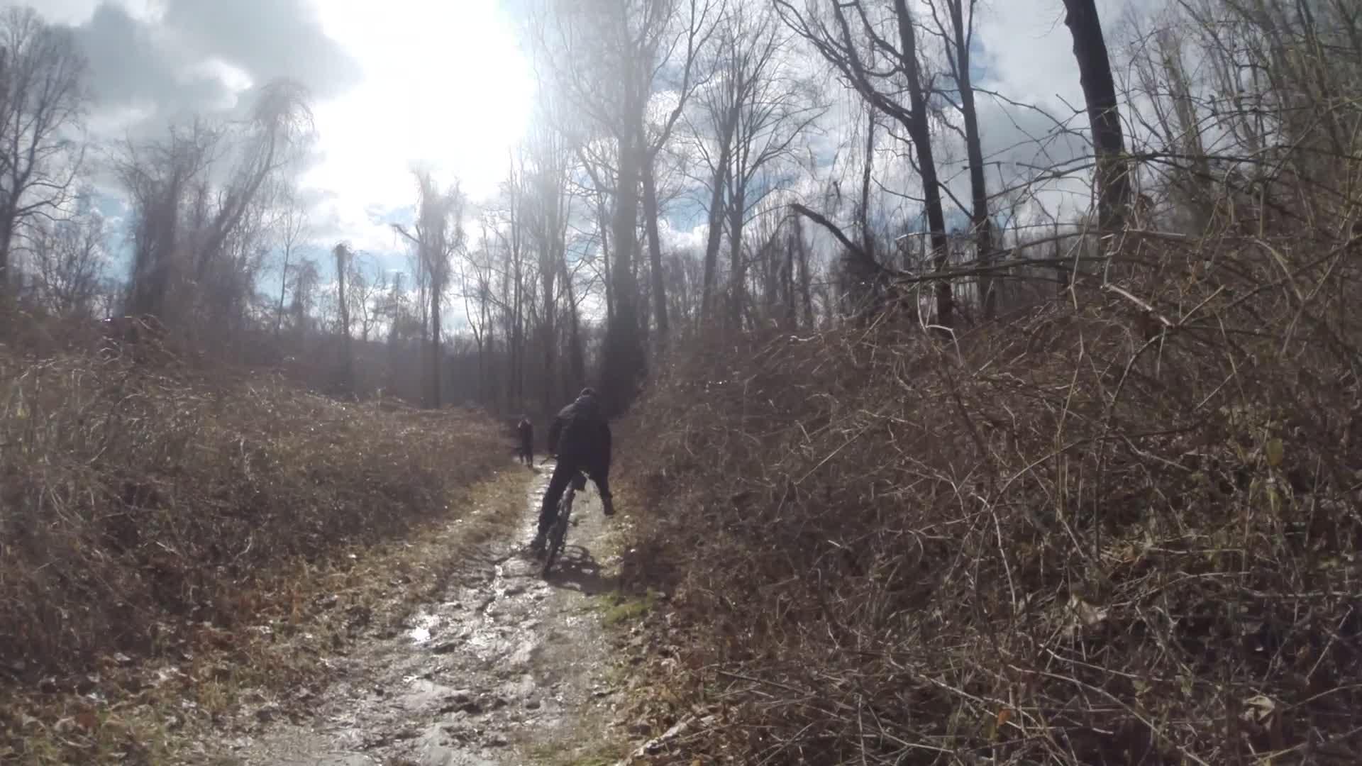 A cyclist riding on a muddy path surrounded by bare trees and brush, with sunlight filtering through the clouds above. Hartshorne Woods Park mountain bike trail.