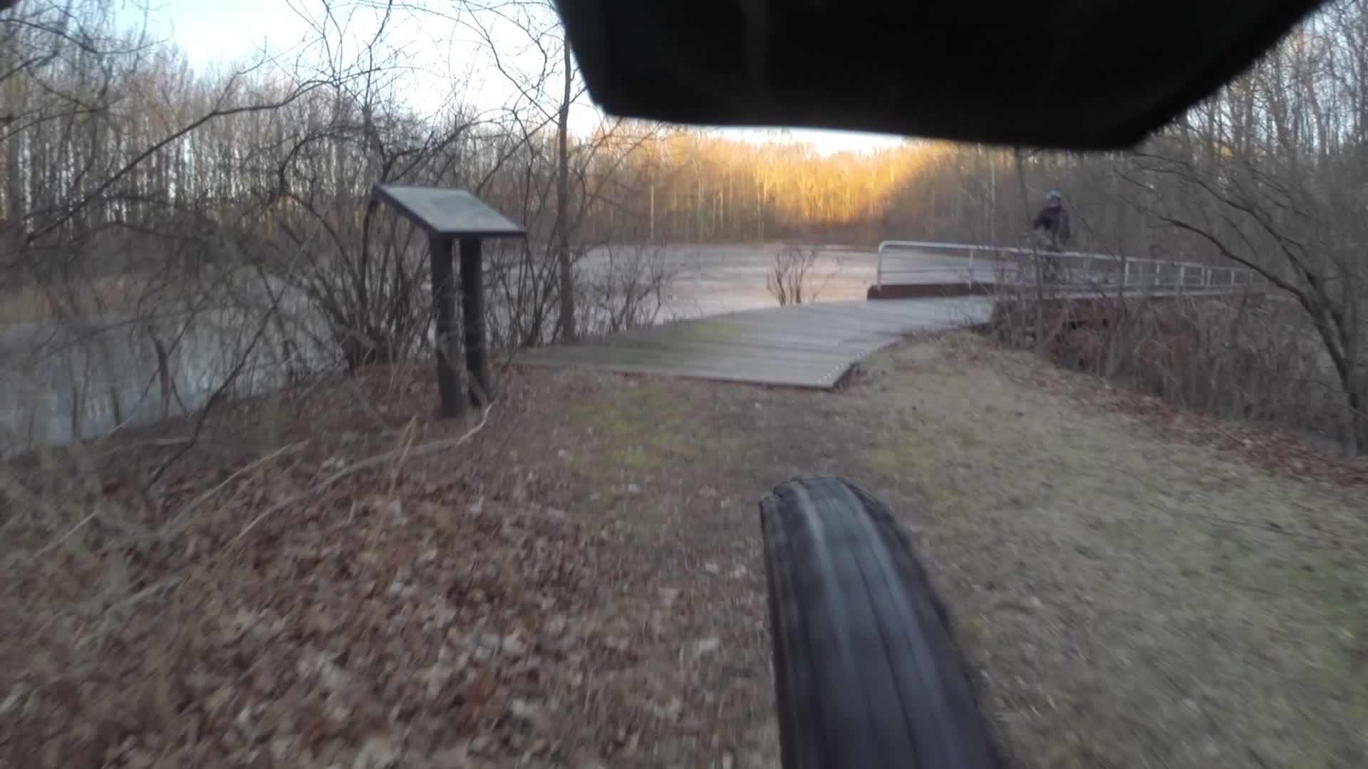 A close-up view of a bicycle tire on a path leading towards a wooden dock by a calm body of water, surrounded by trees. In the background, a person is riding a bike on the dock, with foliage visible around the area during early morning or late afternoon light. A signpost is also present near the water's edge. Trails seperated by streets mountain bike trail.