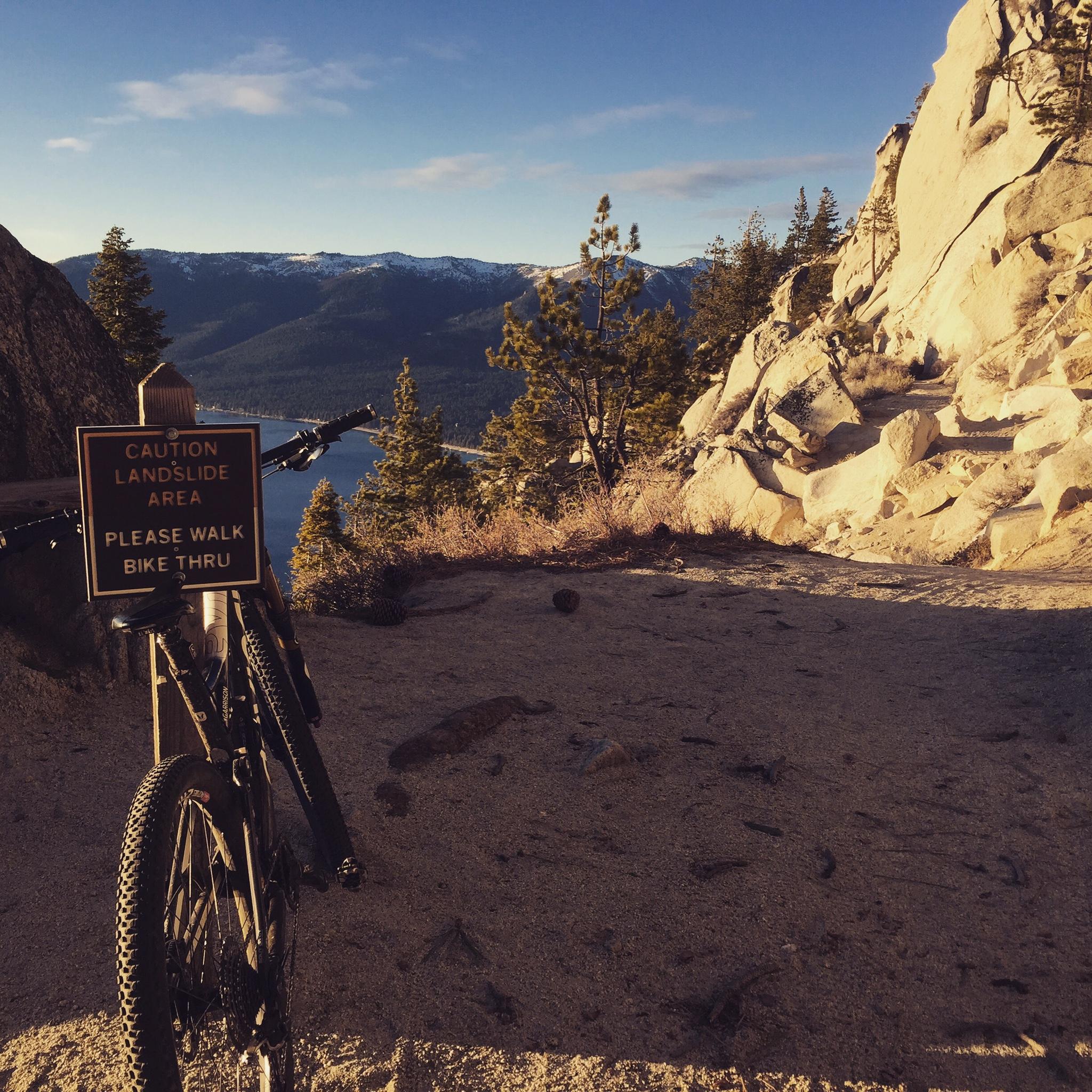 A mountain bike parked beside a caution sign that reads "Caution Landslide Area Please Walk Bike Thru." The background features a scenic view of mountains and a lake under a clear sky, with trees lining the trail. Tahoe Rim Trail: Tahoe Meadows to Tunnel Creek Road / Flume Trail mountain bike trail.