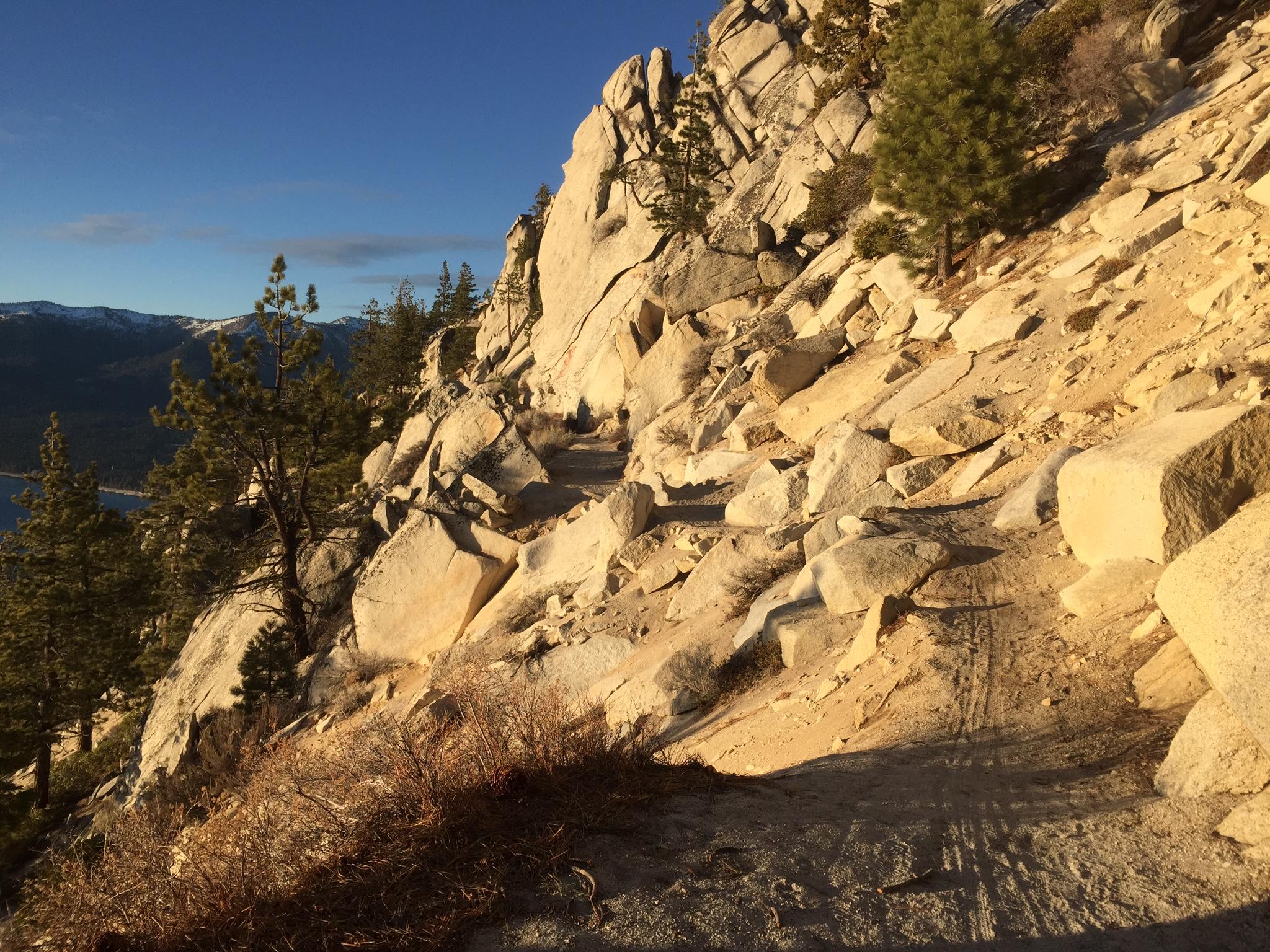 A rocky hiking trail winding along the side of a steep mountain, surrounded by pine trees. The landscape features large boulders and a scenic view of a lake in the distance, with snow-capped mountains under a clear blue sky. The sunlight casts warm tones on the rocky ground. Tahoe Rim Trail: Tahoe Meadows to Tunnel Creek Road / Flume Trail mountain bike trail.