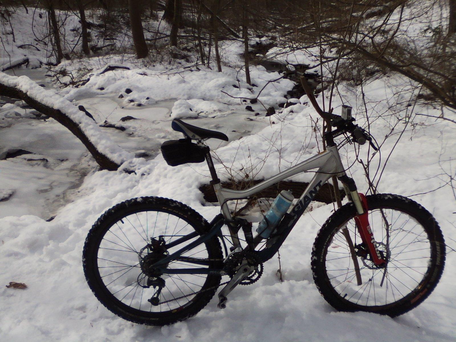 A mountain bike parked beside a frozen creek in a snowy landscape, with trees in the background. The bike features a water bottle holder and is partially covered by snow. Trails seperated by streets mountain bike trail.