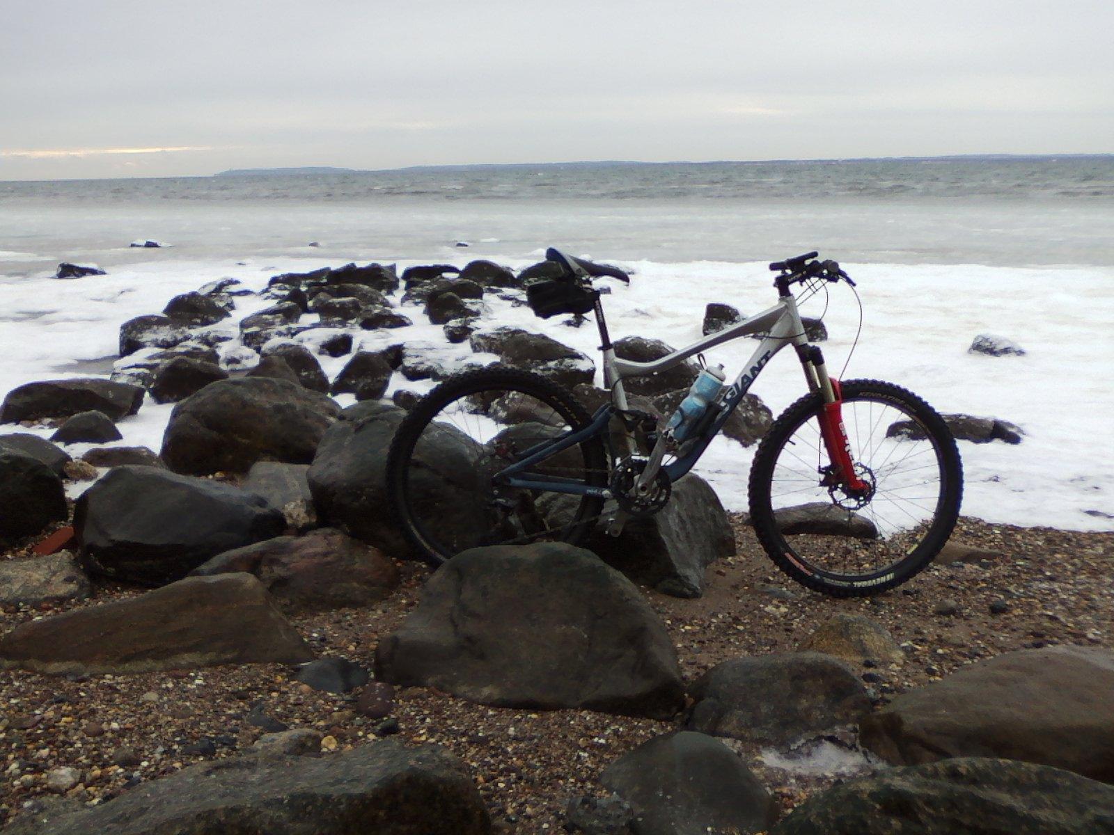 A mountain bike positioned on rocky terrain near the shore, with gentle waves lapping at the rocks. The background shows a cloudy sky and distant land across the water. Wolfes Pond park mountain bike trail.