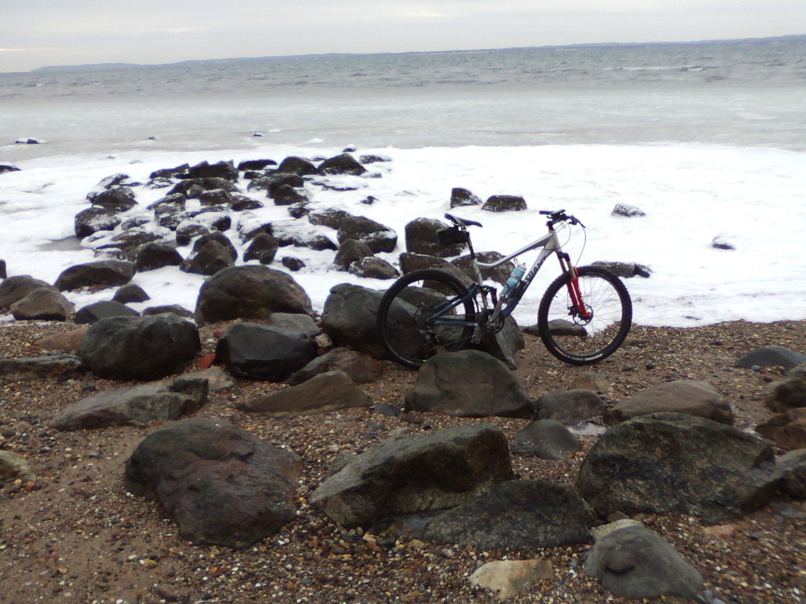 A mountain bike leaning against large rocks on a beach, with waves crashing in the background and a cloudy sky overhead. Wolfes Pond park mountain bike trail.